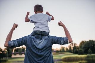 A young father carries his toddler son piggyback as they both raise their fists toward the sky