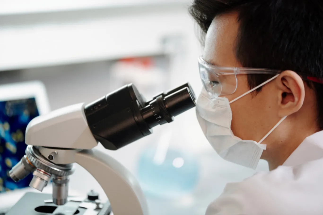 A lab technician wearing a mask looks through a microscope