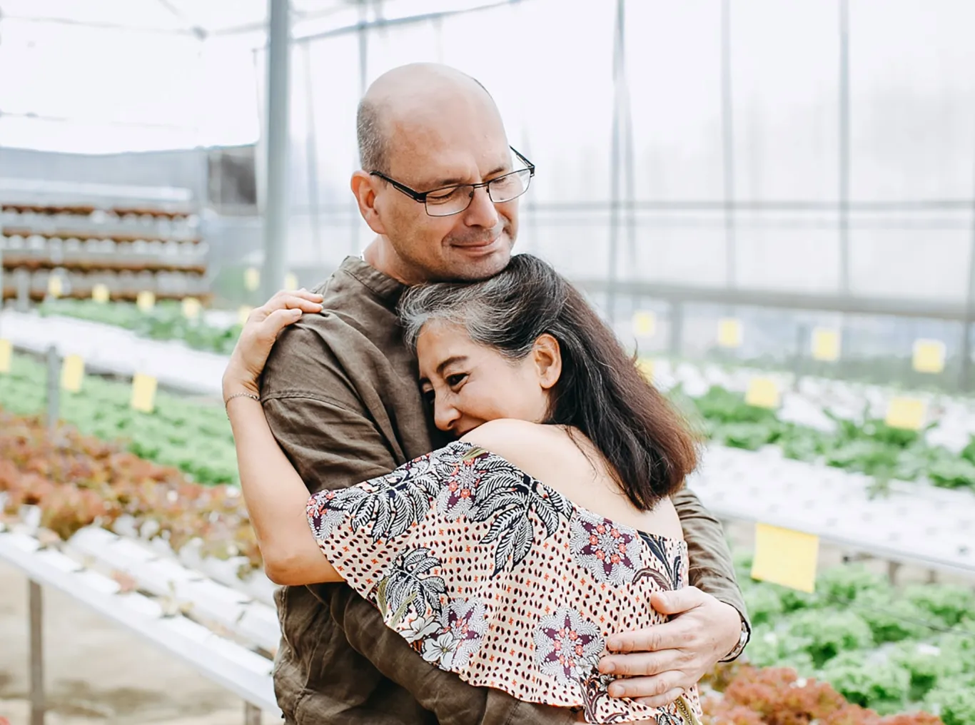 A man hugs his wife as they stand inside a sunlit greenhouse