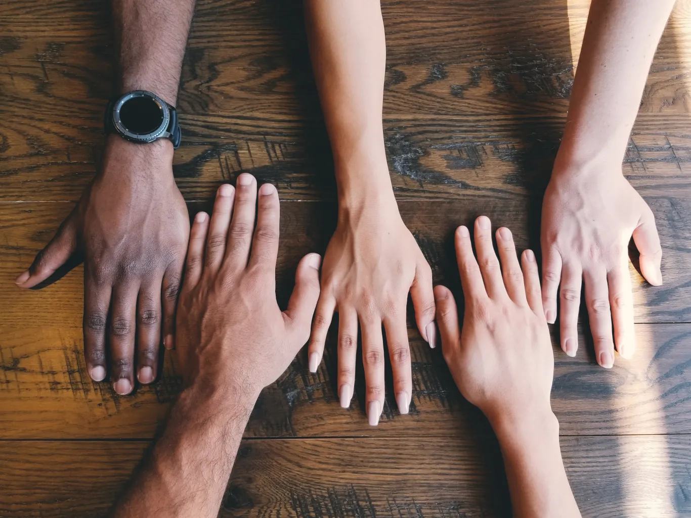 People of diverse races and genders rest their hands beside one another on a wooden table
