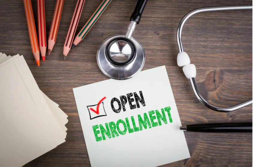An open enrollment sign rests on a desk beside a stethoscope and several colored pencils