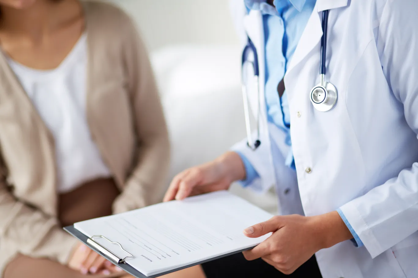 A doctor in a white coat holds a clipboard while talking with a patient