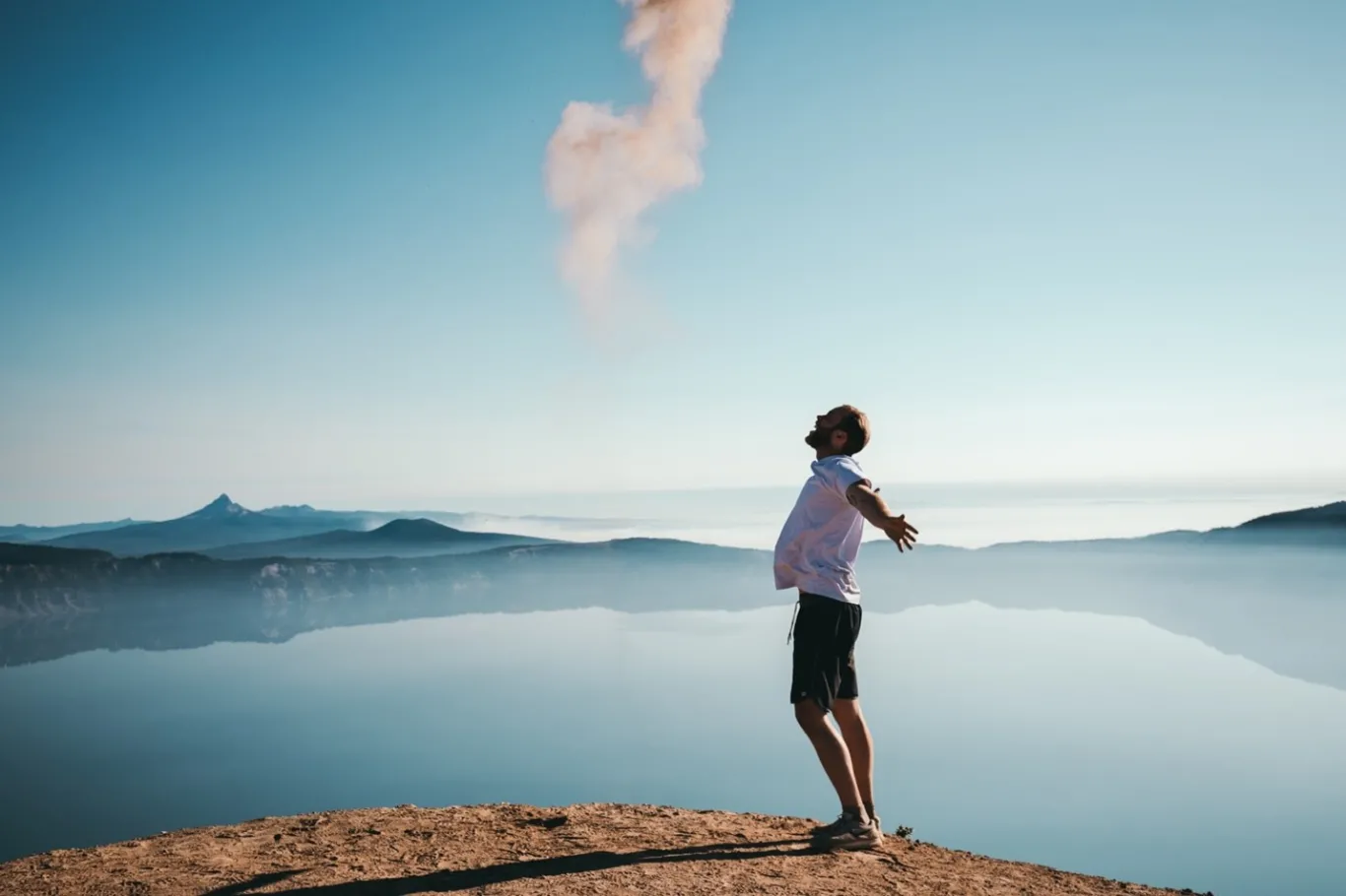 A man in white shirt and shorts stretches out his arms and looks up at the blue sky smiling beside a lake