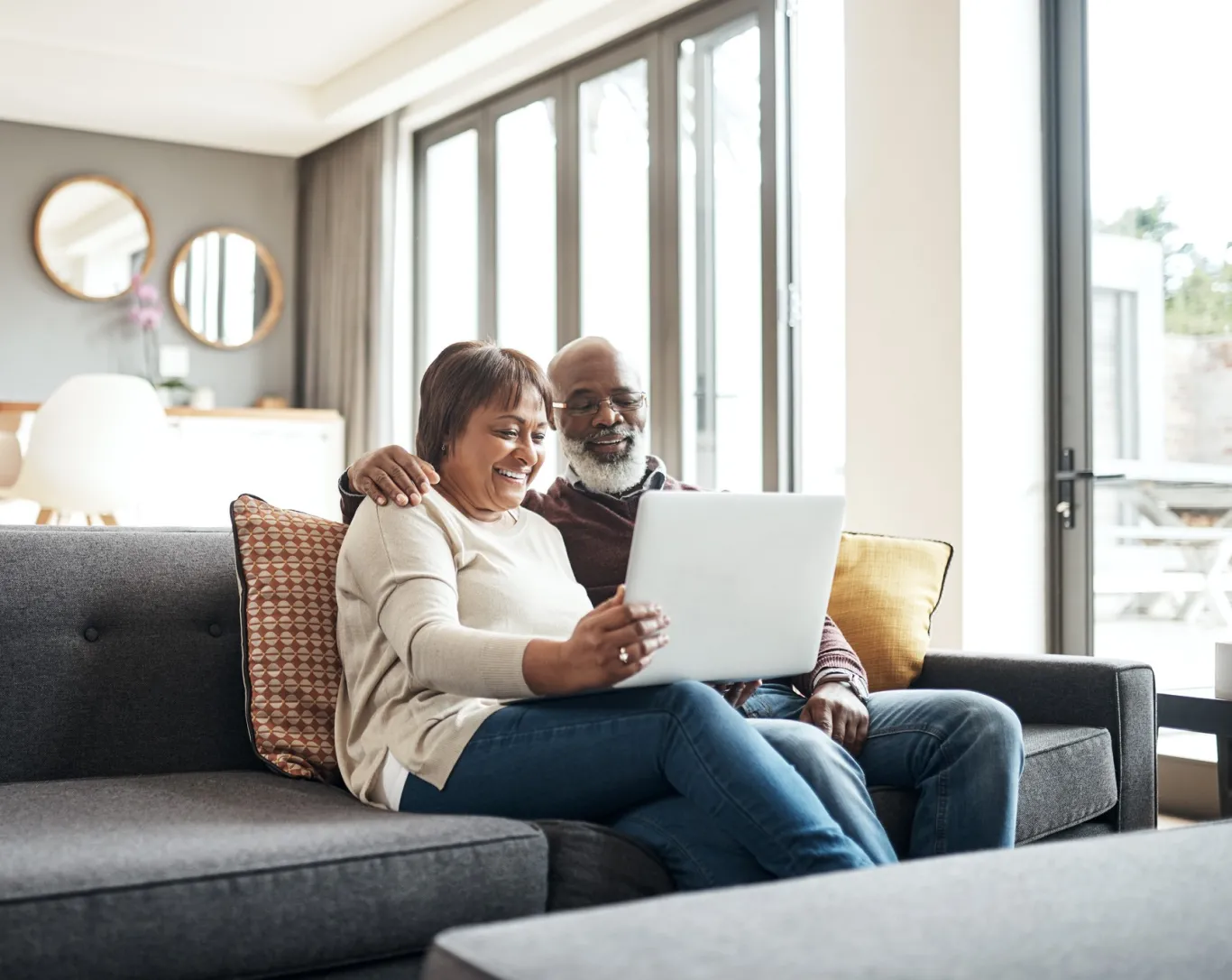A happy couple sits together on their sunlit living room couch looking at their laptop