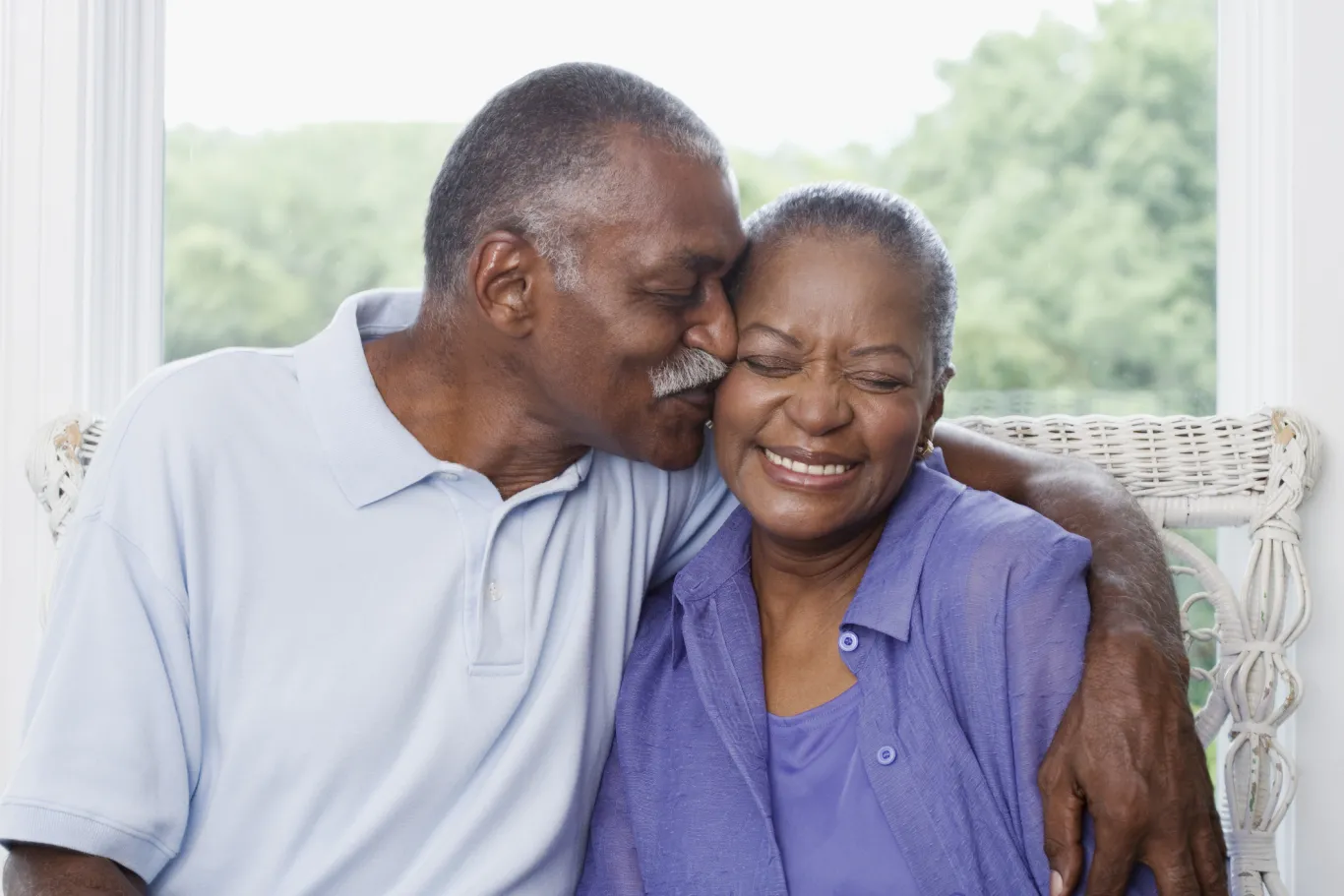 A senior couple smiles and cuddles in their home next to a window overlooking green leafy trees