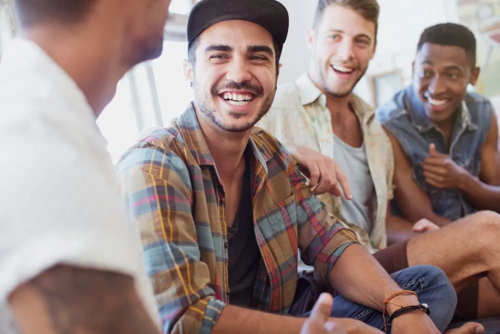 Four young men sit together beside a window laughing and talking together
