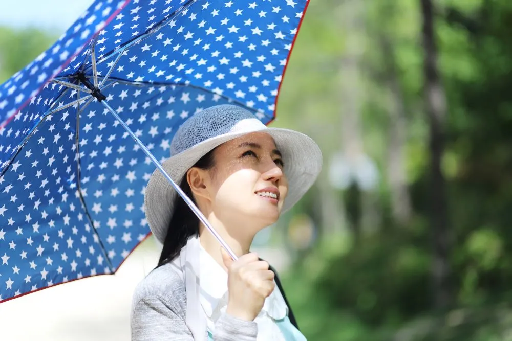 A young woman wears a white hat and carries a blue umbrella to shield her face from the sun