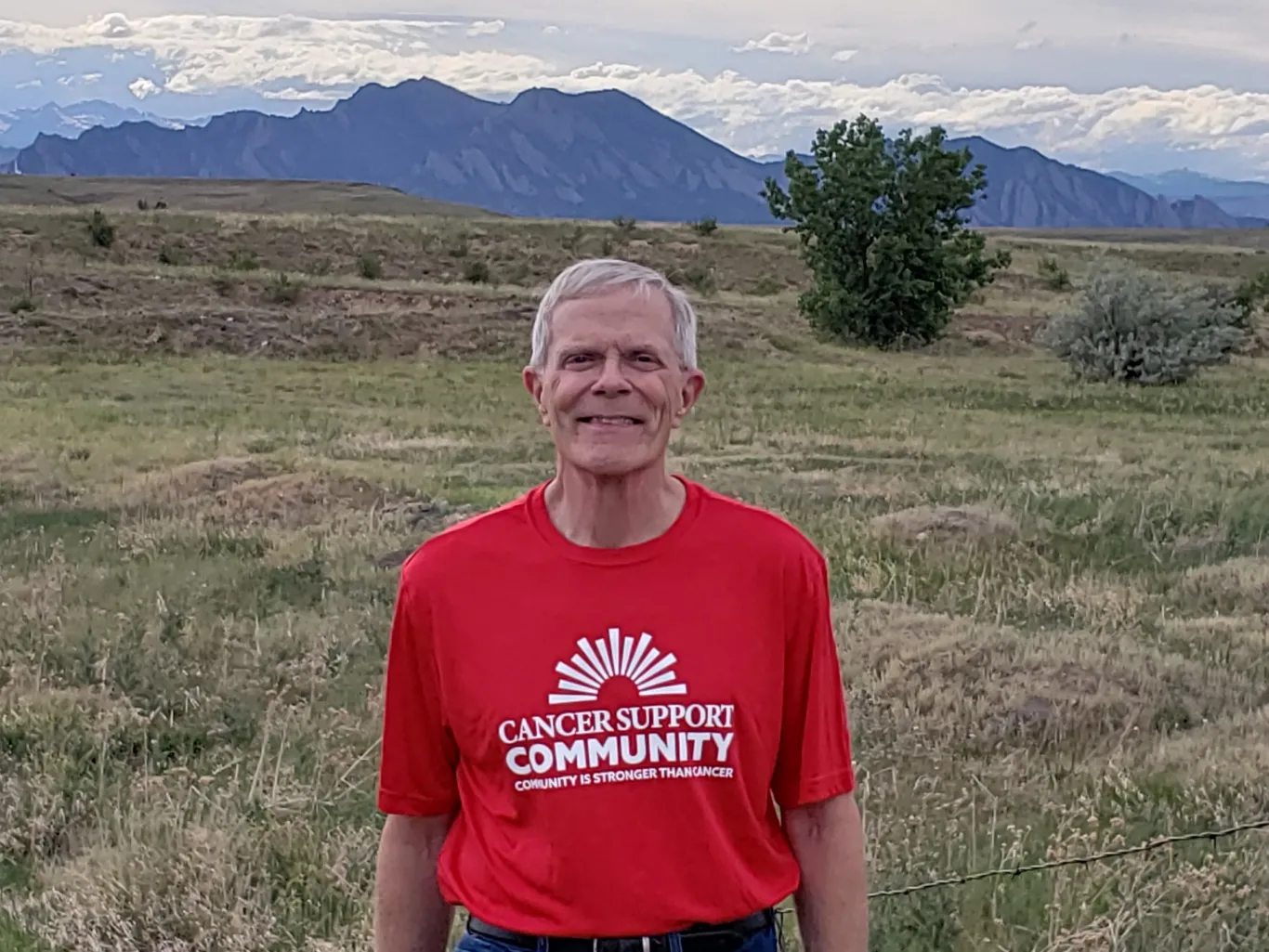 Dressed in jeans and a red Cancer Support Community T-shirt, Paul smiles outside with the Boulder foothills behind him