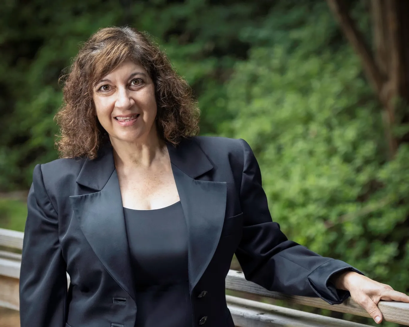 Kerri Susko wears a black business suit and smiles from a wooden bridge beside trees