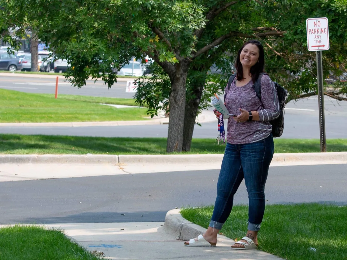 Amber smiles while standing on a sidewalk in front of a leafy green tree in the summer sunshine 