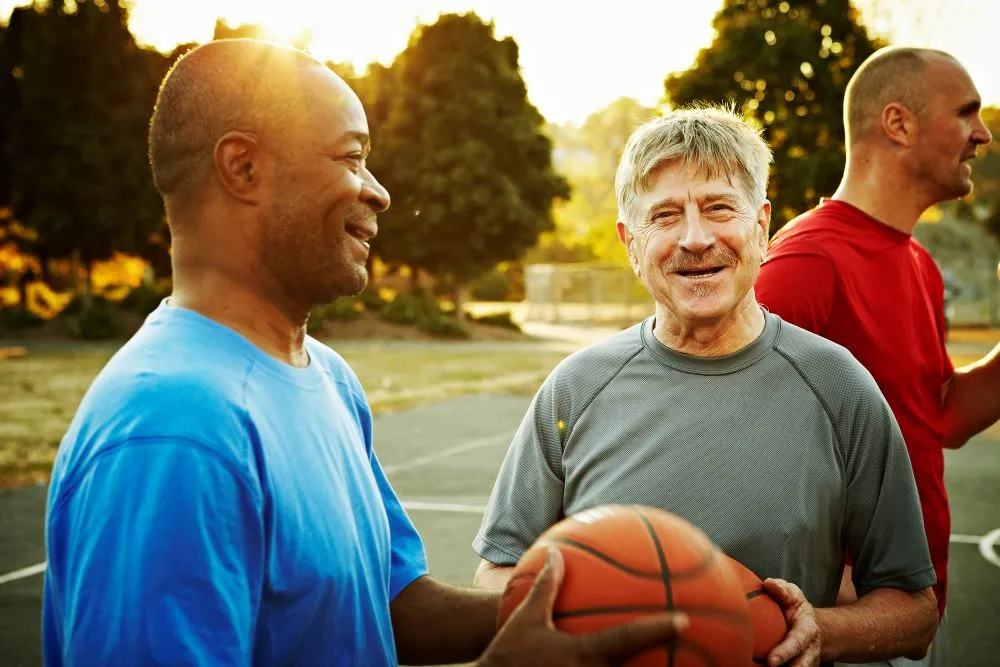 Three men in athletic clothing stand together on a sunlit basketball court.