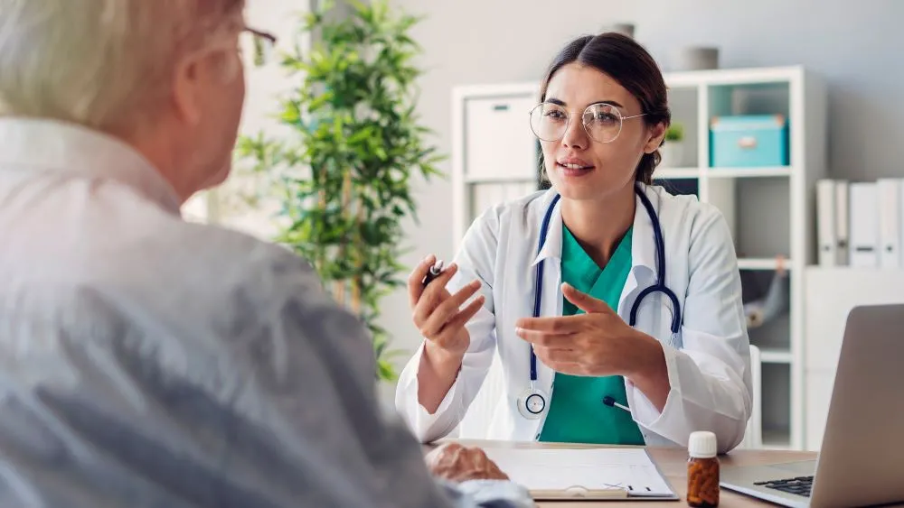 A woman doctor in a white lab coat speaks with a man sitting at a desk in a doctor's office