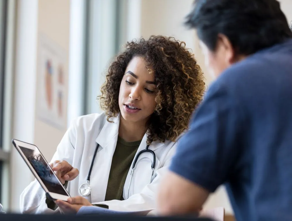 A female doctor in a white coat shows a male patient information on a tablet screen