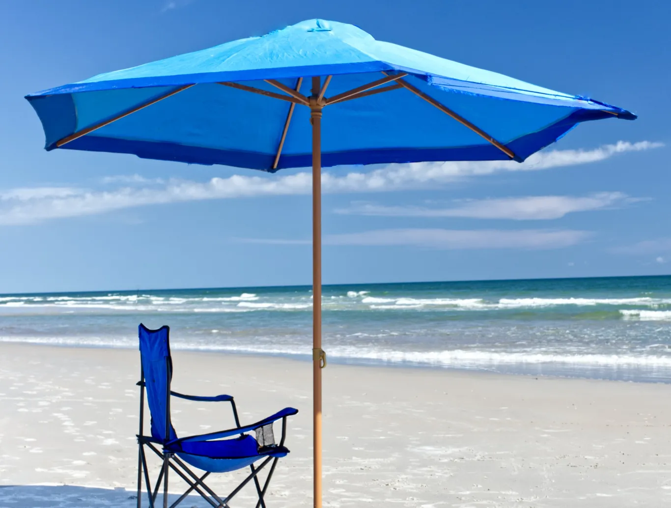 A blue beach umbrella casts shade over a blue folding chair on a sunny beach