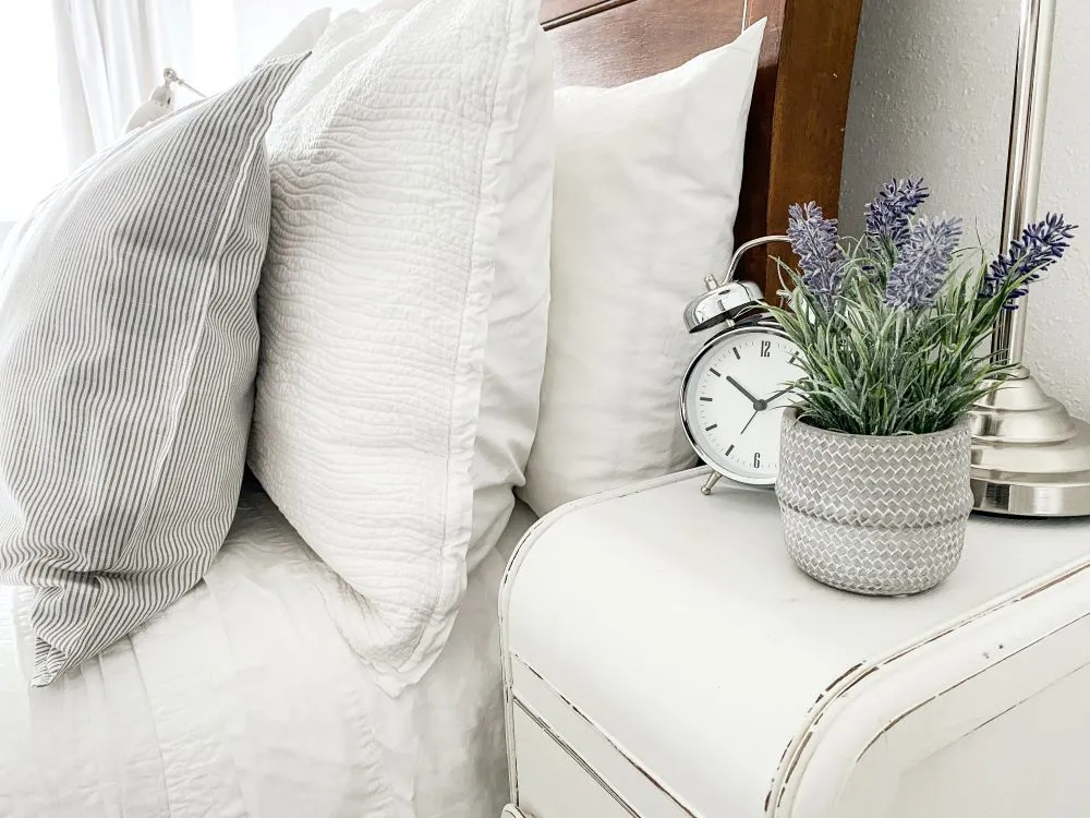 Pillows are stacked on a bed next to a table with alarm clock and potted lavender flowers