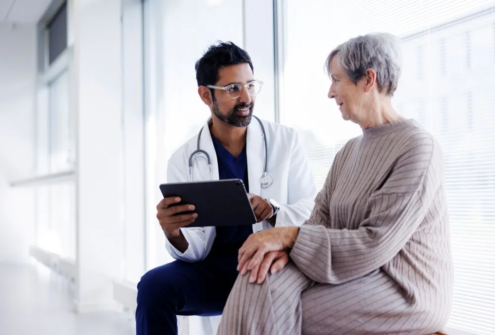 A woman with short gray hair talks with a doctor in a lab coat in a brightly lit room