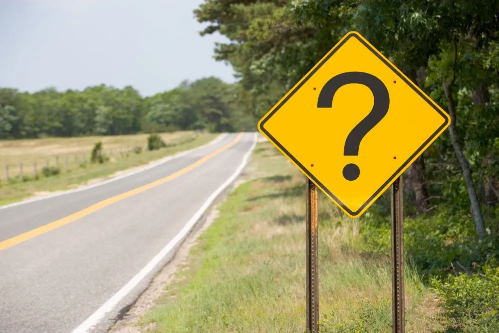 A yellow road sign on the side of an empty road displays a large question mark
