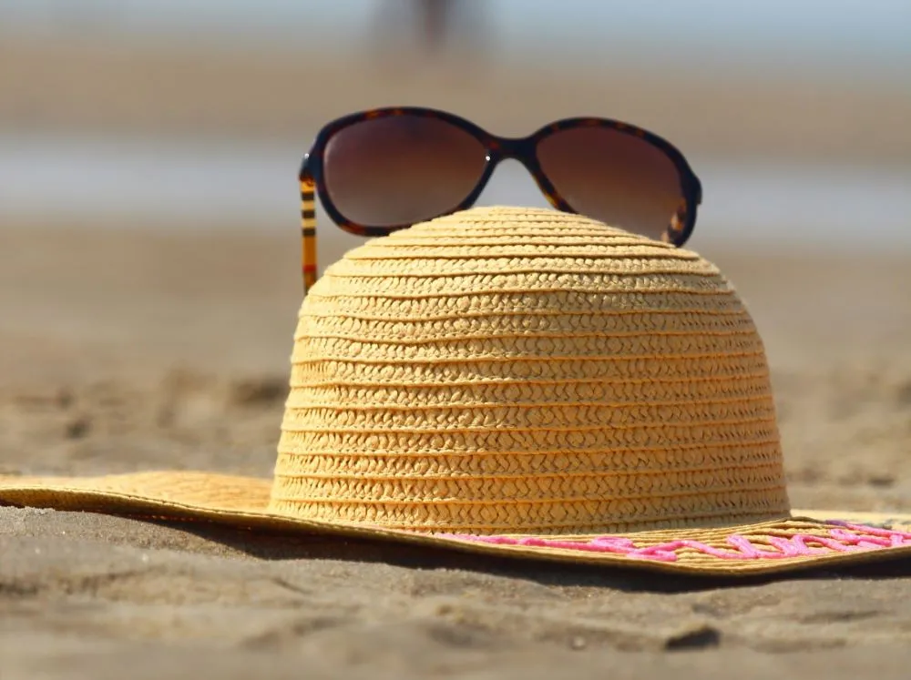 A pair of brown sunglasses sits on a straw sun hat resting on a sandy beach