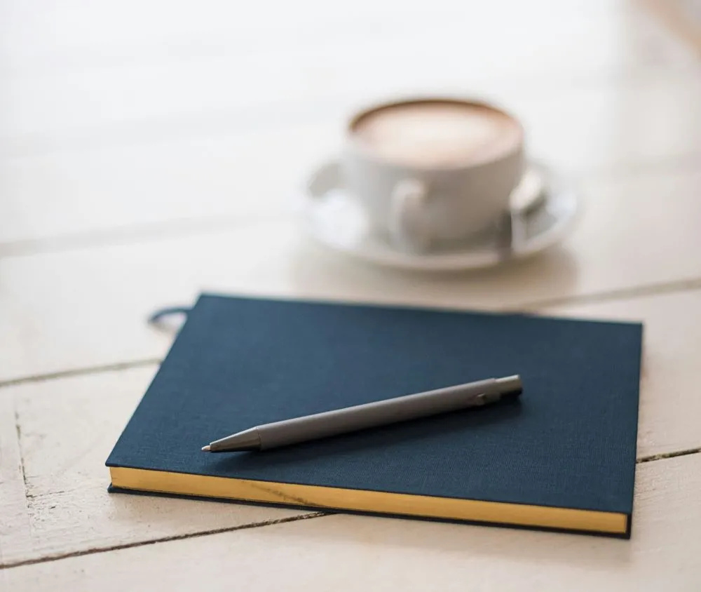 A black pen rests on a hard-cover notebook beside a cup of coffee
