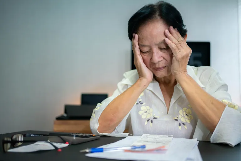 A distressed elderly woman rests her face in her hands as she looks at some bills on her desk