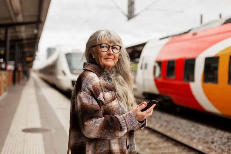 A mature woman with long hair and dark glasses waits at a train station