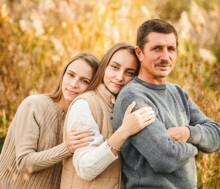 A father and his daughters stand beside one another in front of a sunlit field of grass
