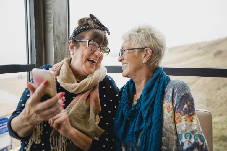 Two women friends laugh together beside a window while looking at their smartphone