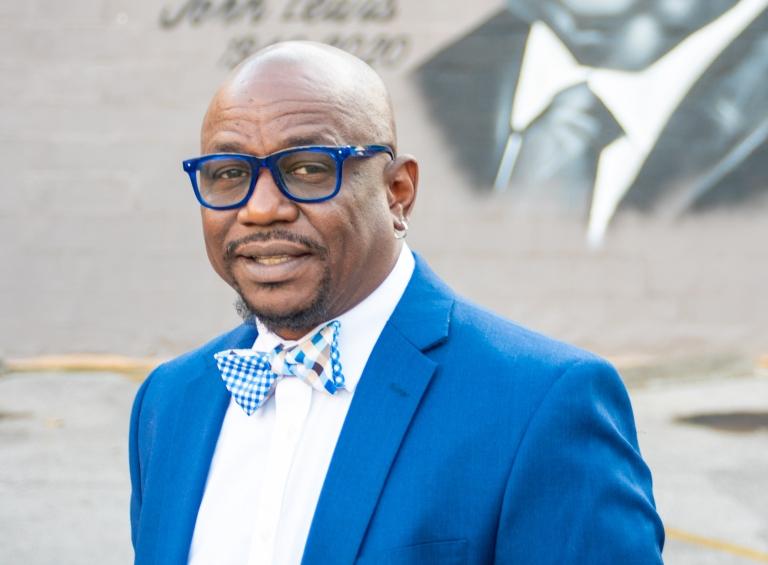 Tony Walker wears a blue and white bow tie, blue blazer, white blouse, and glasses with blue frames