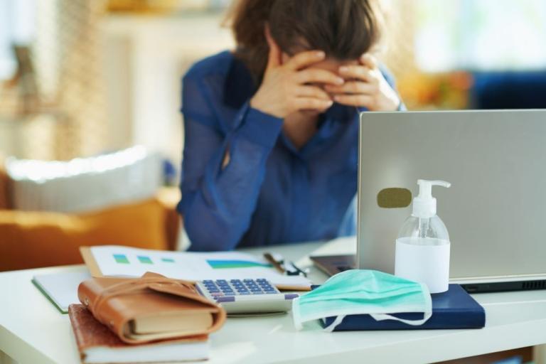 A woman in a blue blouse rests her face in her hands at a desk with a laptop, calculator, and worksheets