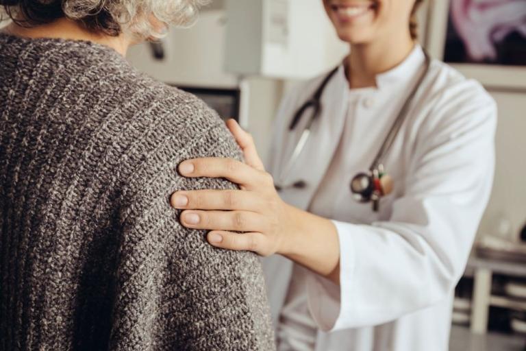A female doctor in a white lab coat rests a comforting hand on a patient's shoulder