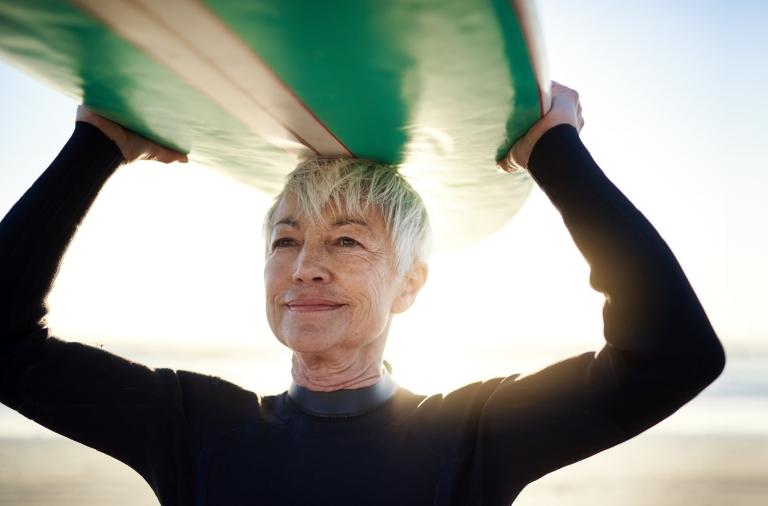 A senior woman with short gray hair holds a surf board over her head on a sunny beach