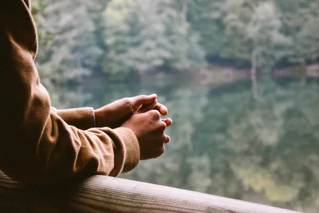 An unseen person rests their hands on wood bridge looking out at a reflective tree-lined lake