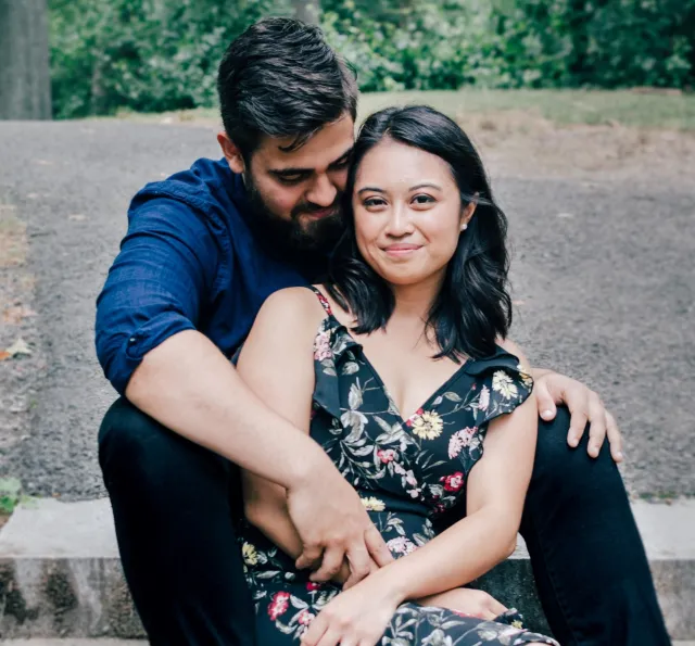 A smiling couple cuddles together outdoors on a tree-lined trail