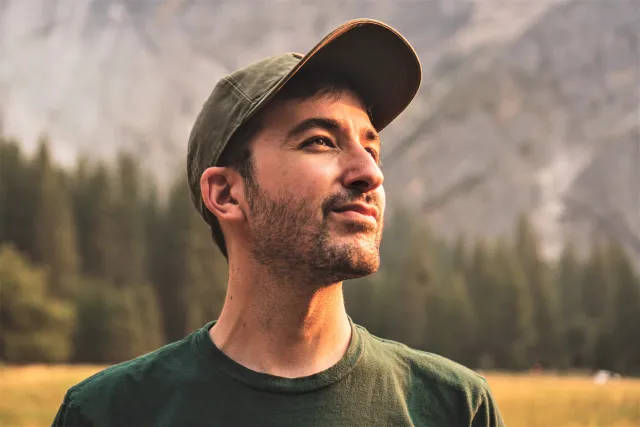 A young man in a ballcap stares up at the sky with mountains in the background