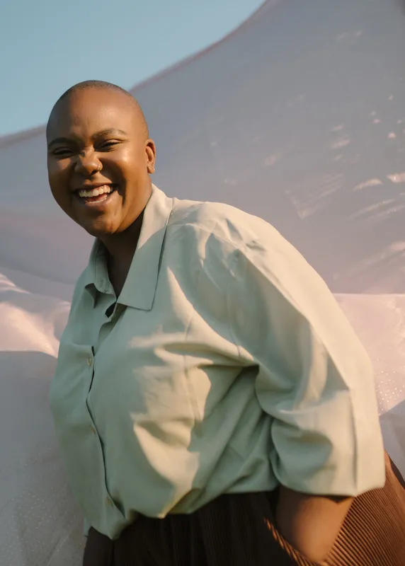 Bald woman laughs with her hand in her pocket and sun on her face.