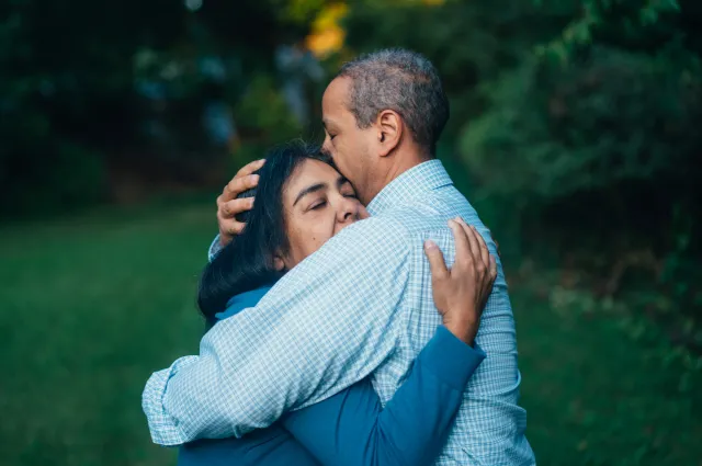 A man hugs his wife outdoors in a tree-lined yard