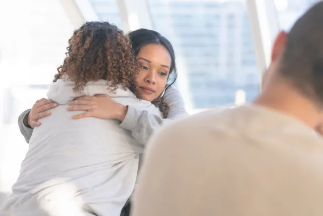 Two (2) women embrace in a comforting hug.