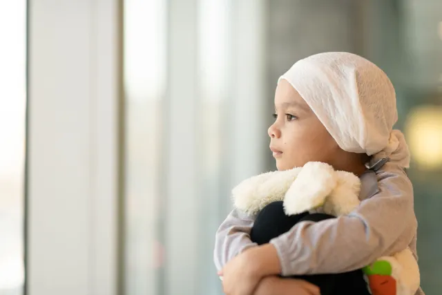 A young girl wearing a headscarf holding a stuffed animal looking out a window
