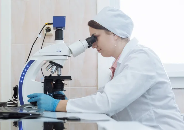A lab technician wearing a white coat and cap peers into a microscope sitting on a reflective table