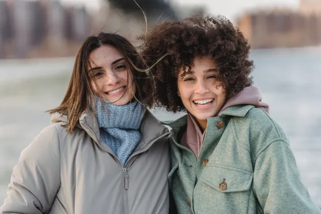 Two young women wearing winter coats smile together outdoors in a snowy field