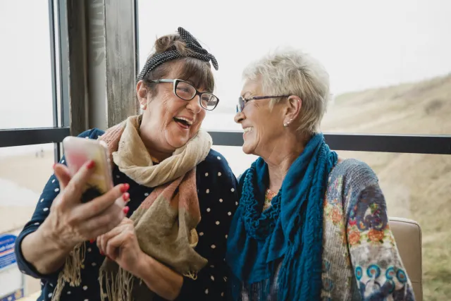 Two women friends laugh together beside a window while looking at their smartphone