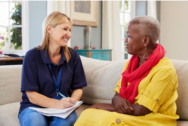A woman in a red scarf and yellow dress sits and chats with her social worker.