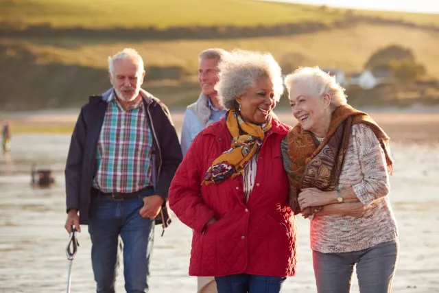 A group of smiling senior friends wearing scarves and jackets walks along a breezy shoreline