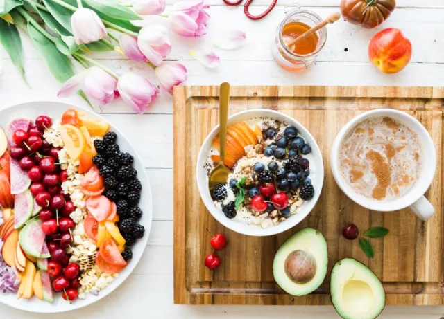 A breakfast displays fresh fruits sits alongside a wood cutting board with a bowl of cereal, avocado, and a cup of tea