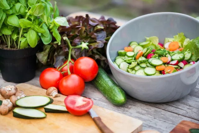 A wooden table displays basil, lettuce leaves, mushrooms, and bright red tomatoes in a sunny outdoor setting