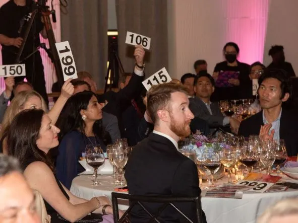 People seated at an elegant dining table hold up their bids for bottles of champagne during CSC's Annual Celebration