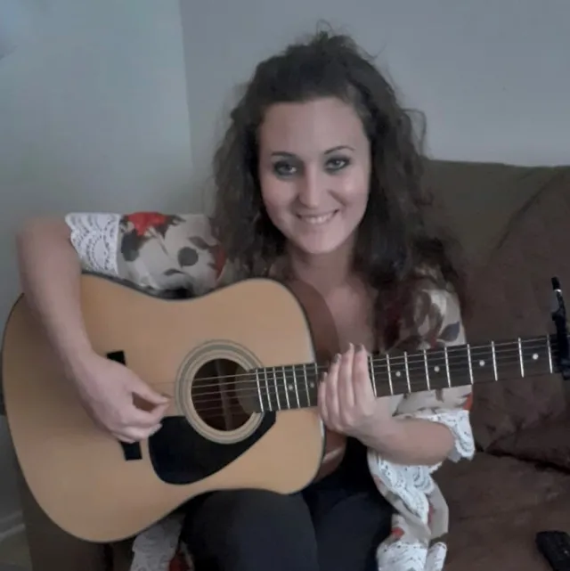 Seated on a couch, Kristen smiles while plucking a guitar that rests in her lap