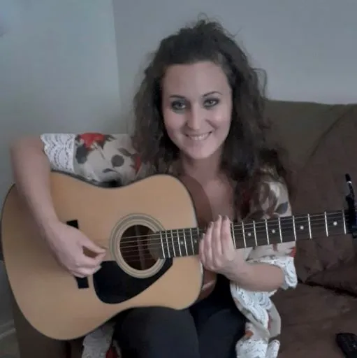 Seated on a couch, Kristen smiles while plucking a guitar that rests in her lap