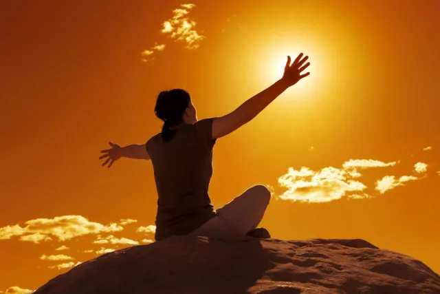 A woman sitting on a rock extends her arms out as if embracing the setting sun in an orange-lit sky