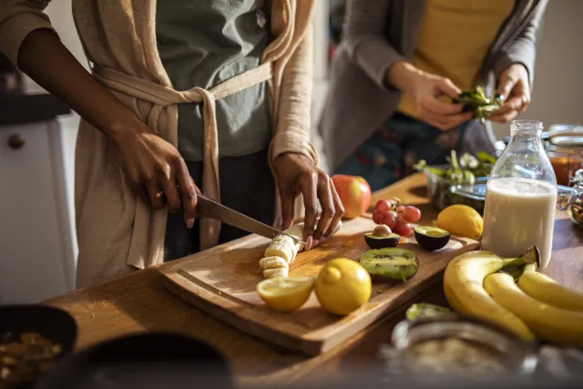 Women prepare veggies and fruits on a cutting board in a brightly lit kitchen